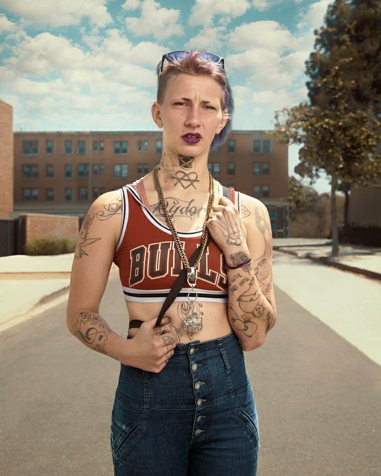 Portrait of a slender tattooed woman in a Chicago Bulls crop top and denim jeans stands on a city street, brick building in background.